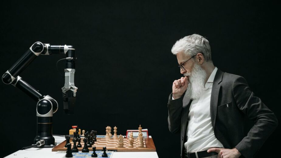 An elderly scientist contemplates a chess move against a robotic arm on a chessboard.