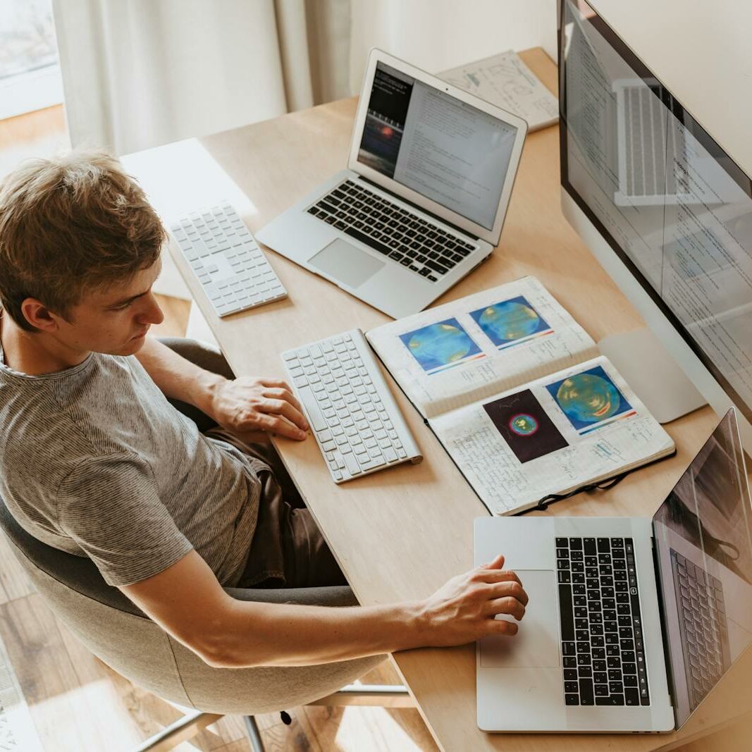 A man multitasks with laptops and a desktop, coding in a home office setting.